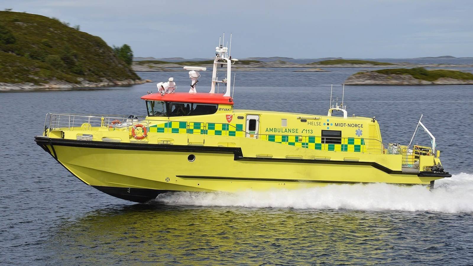 A bright yellow ambulance boat running at speed on open water, marked with green chequer along the hull side and the word AMBULANSE, her red-roofed wheelhouse forward and crew on deck.