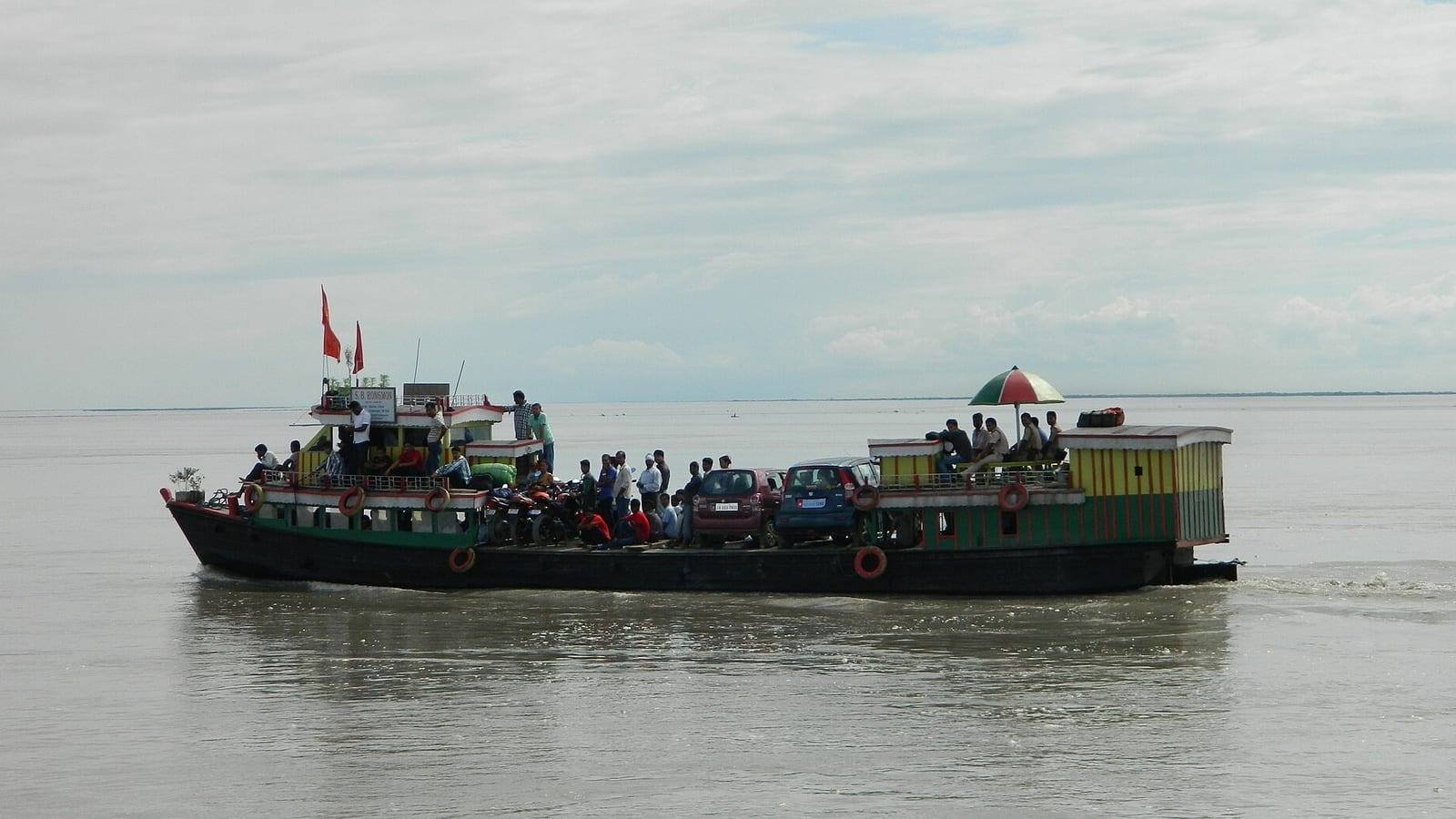 A loaded river ferry under way on a wide, flat river, her open deck carrying cars, motorcycles and a standing crowd of passengers, with flags at the mast and lifebuoys along the bulwark.
