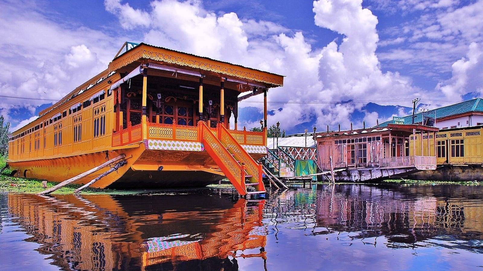 An ornately carved timber houseboat moored on still water, its orange and yellow painted front and open verandah reflected below, with more houseboats along the bank behind.