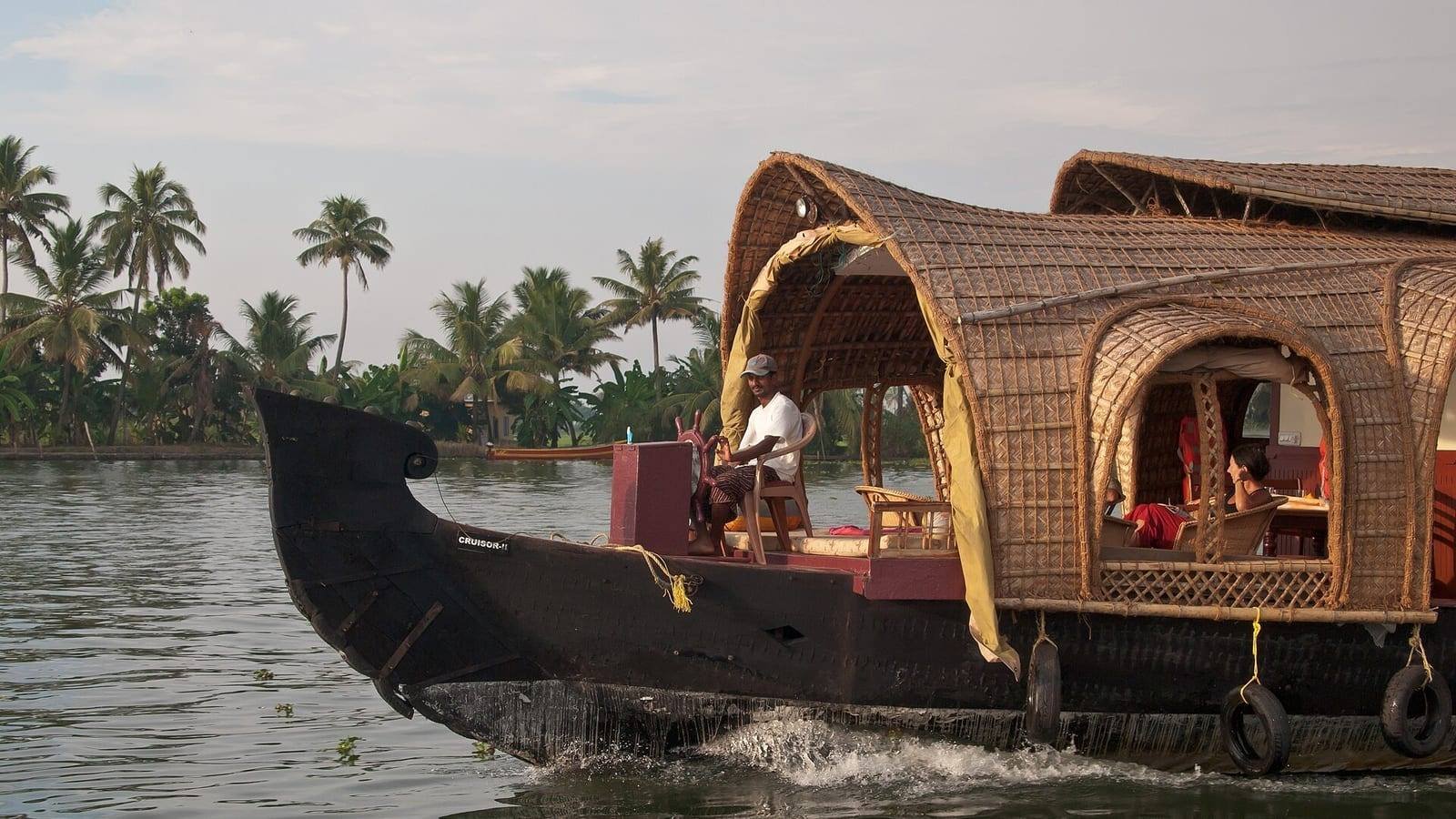 A traditional Kerala houseboat under way on a palm-fringed backwater, her black timber hull curving up at the bow and a woven cane superstructure over the after half, with the boatman standing at the helm.