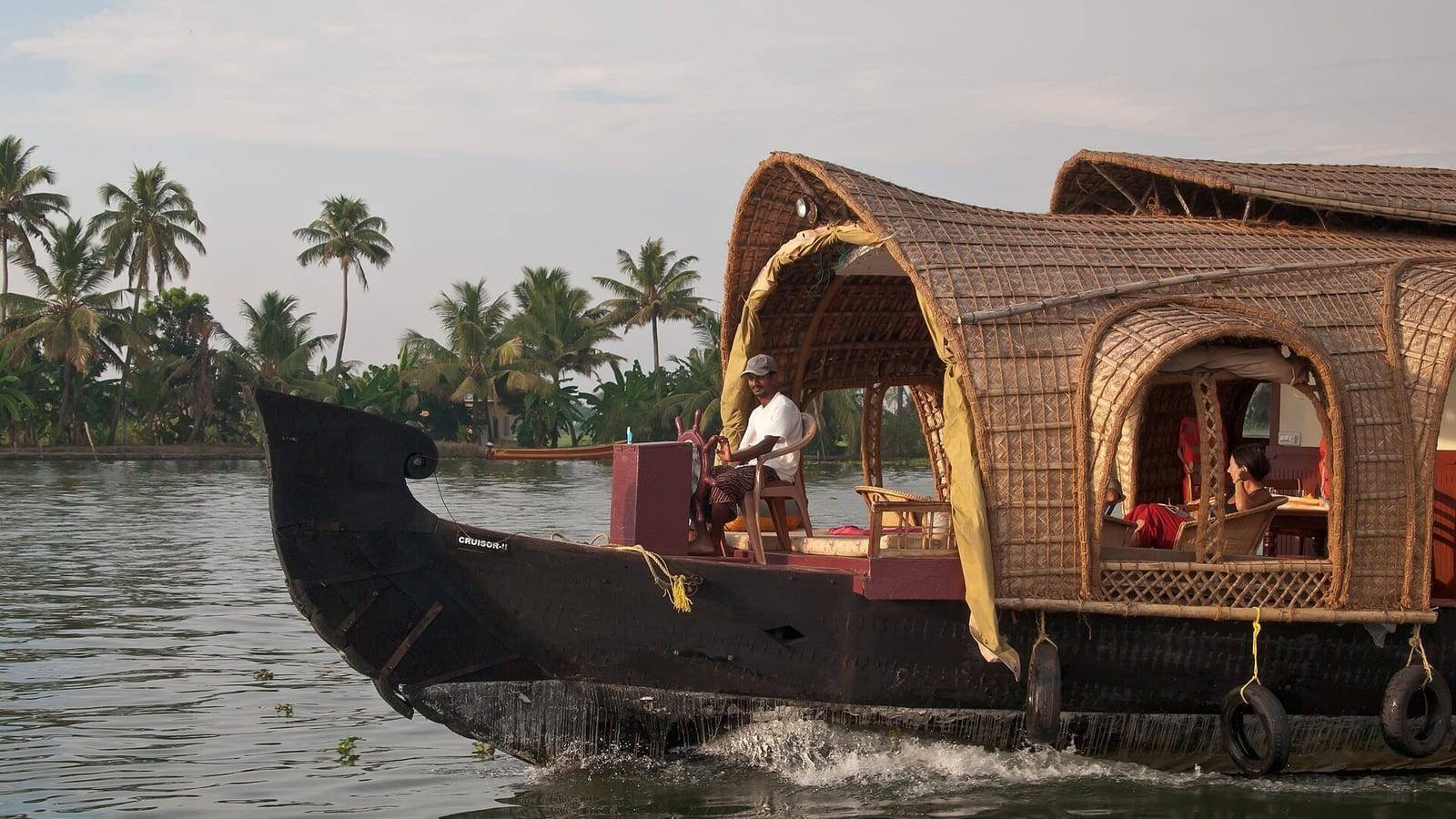 A traditional Kerala houseboat under way on a palm-fringed backwater, her black timber hull curving up at the bow and a woven cane superstructure over the after half, with the boatman standing at the helm.