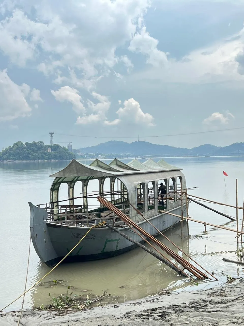 A vessel afloat at the riverbank with her arched canopy fitted and bamboo staging still alongside, hills rising on the far bank.