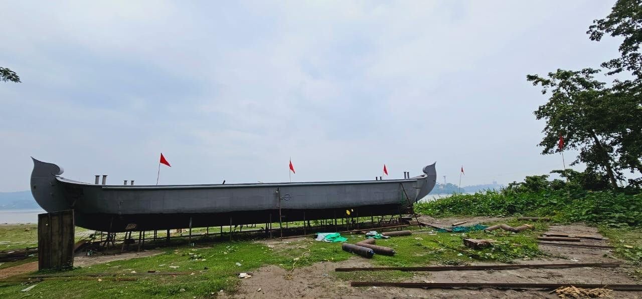A bare steel hull standing on timber blocks on the riverbank, seen side-on along its full length, with upswept bow and stern, marker flags along her sheer and gas cylinders and steel stock lying on the ground alongside.