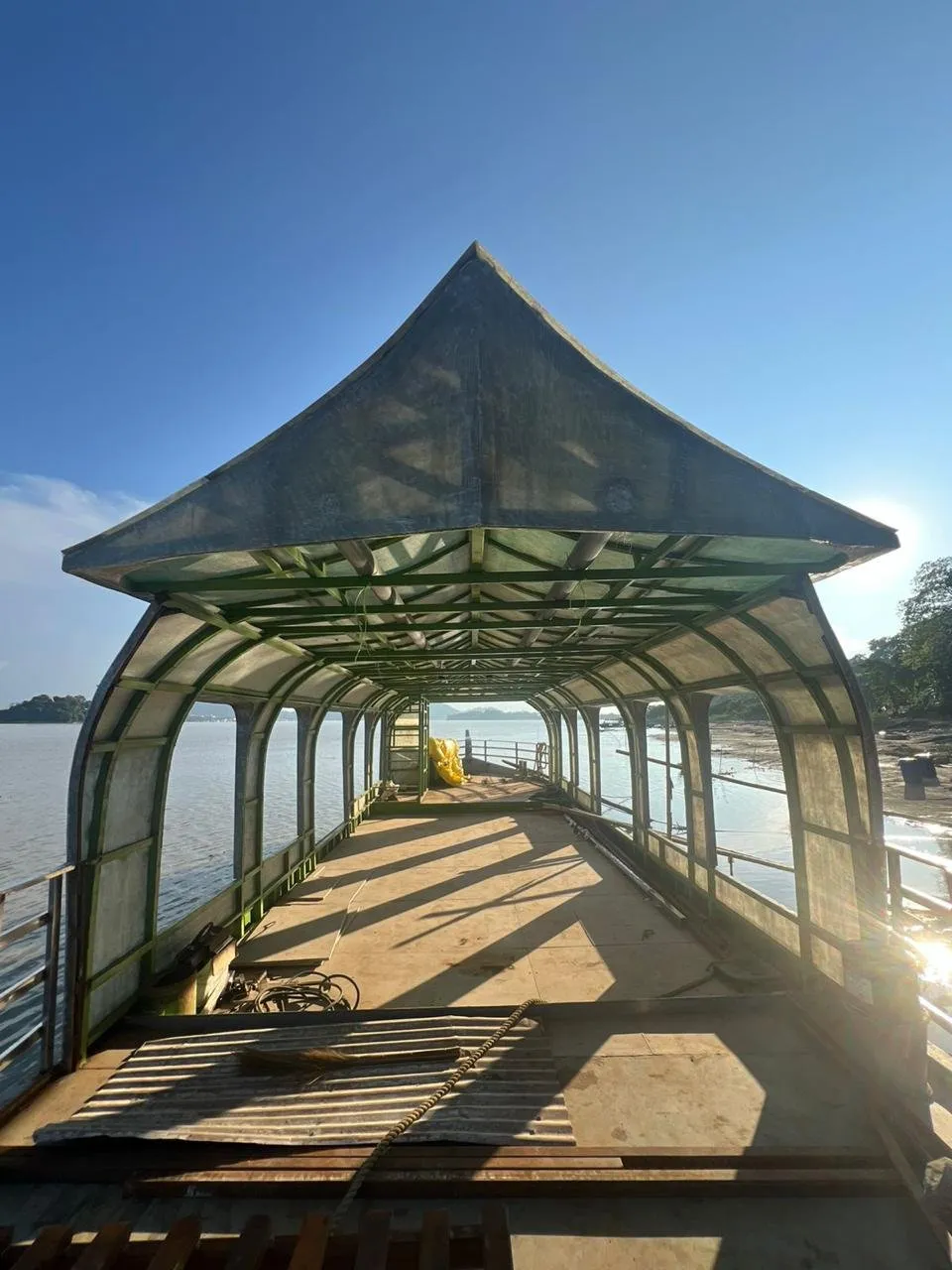 Looking down the open deck of a vessel under construction, beneath her arched canopy and peaked roof, with the river on both sides.