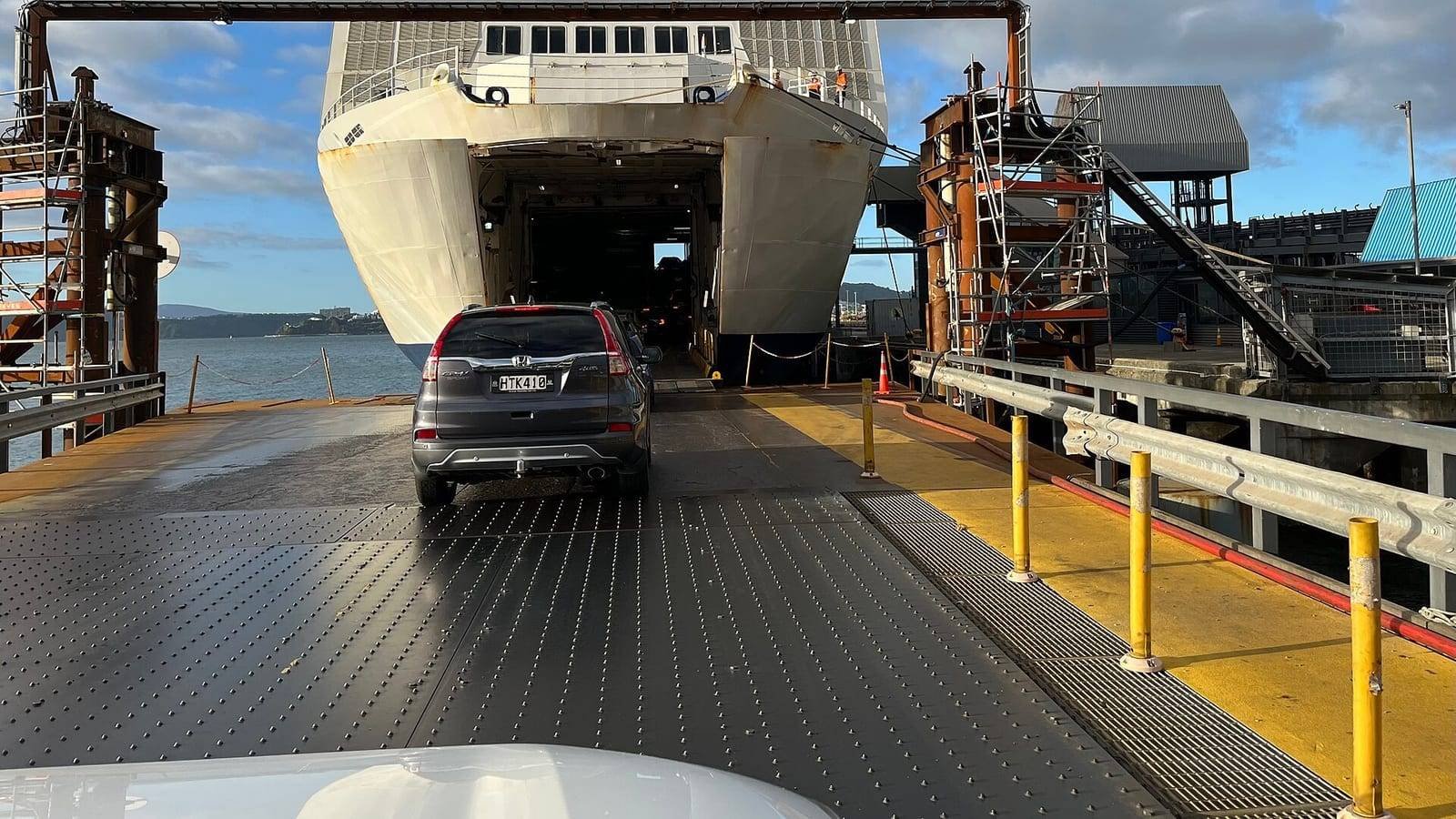 A car driving up a steel linkspan into the open bow of a roll-on roll-off ferry, the vessel’s white superstructure and orange-topped bridge above the vehicle opening.