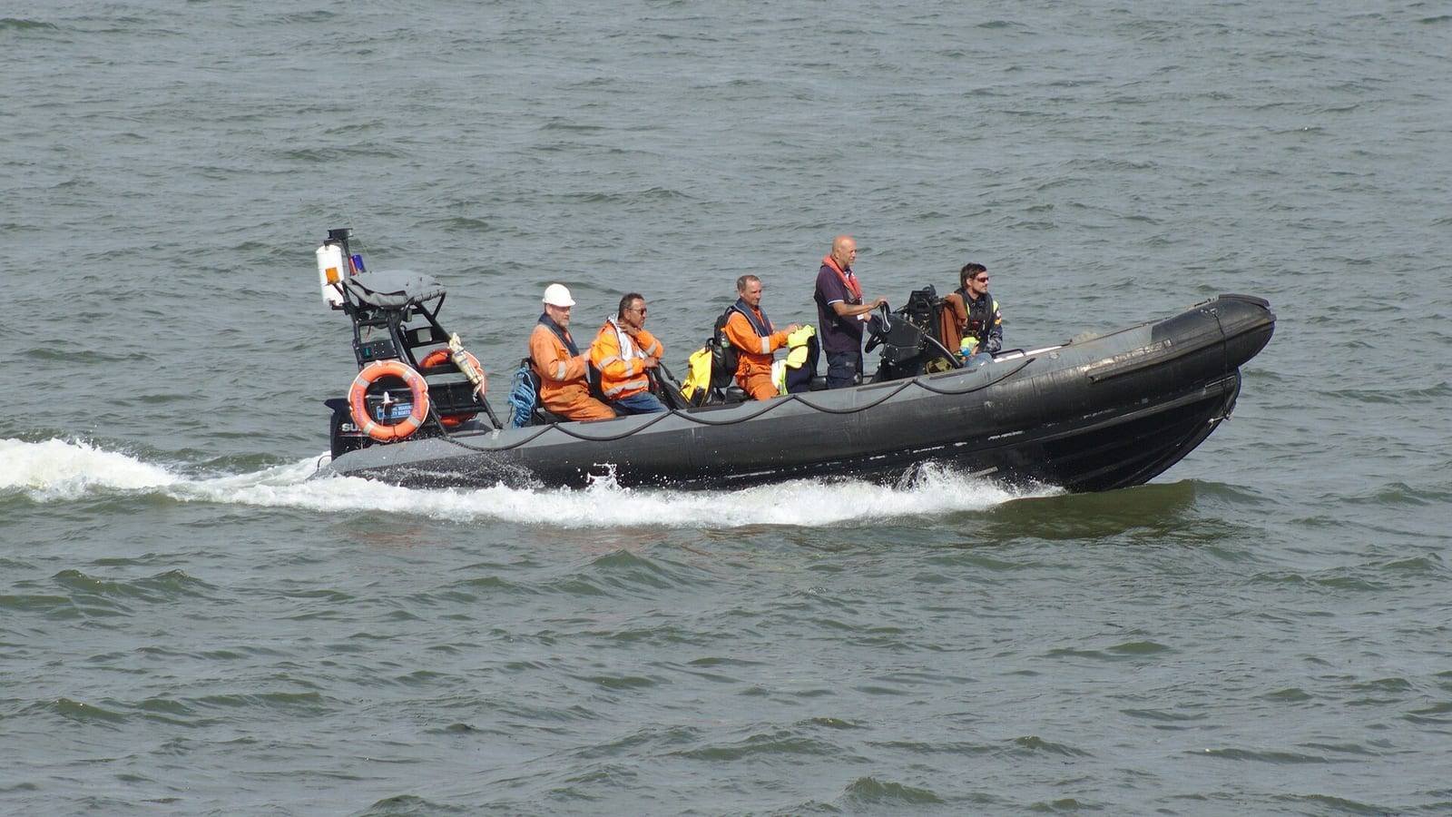 A black rigid inflatable boat under way on choppy grey water with five crew aboard in orange high-visibility gear and helmets, an orange lifebuoy stowed on the foredeck.