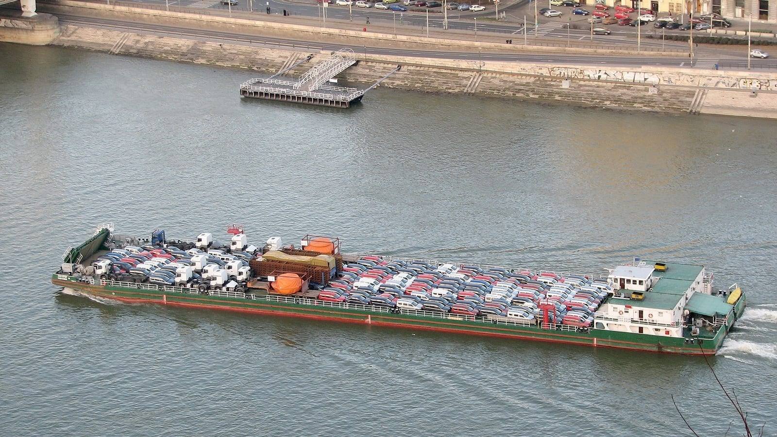 A self-propelled river barge under way, her long open hold packed with new cars and her white wheelhouse aft, seen from above with the far bank behind.