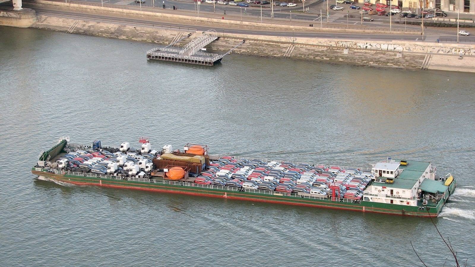 A self-propelled river barge under way, her long open hold packed with new cars and her white wheelhouse aft, seen from above with the far bank behind.