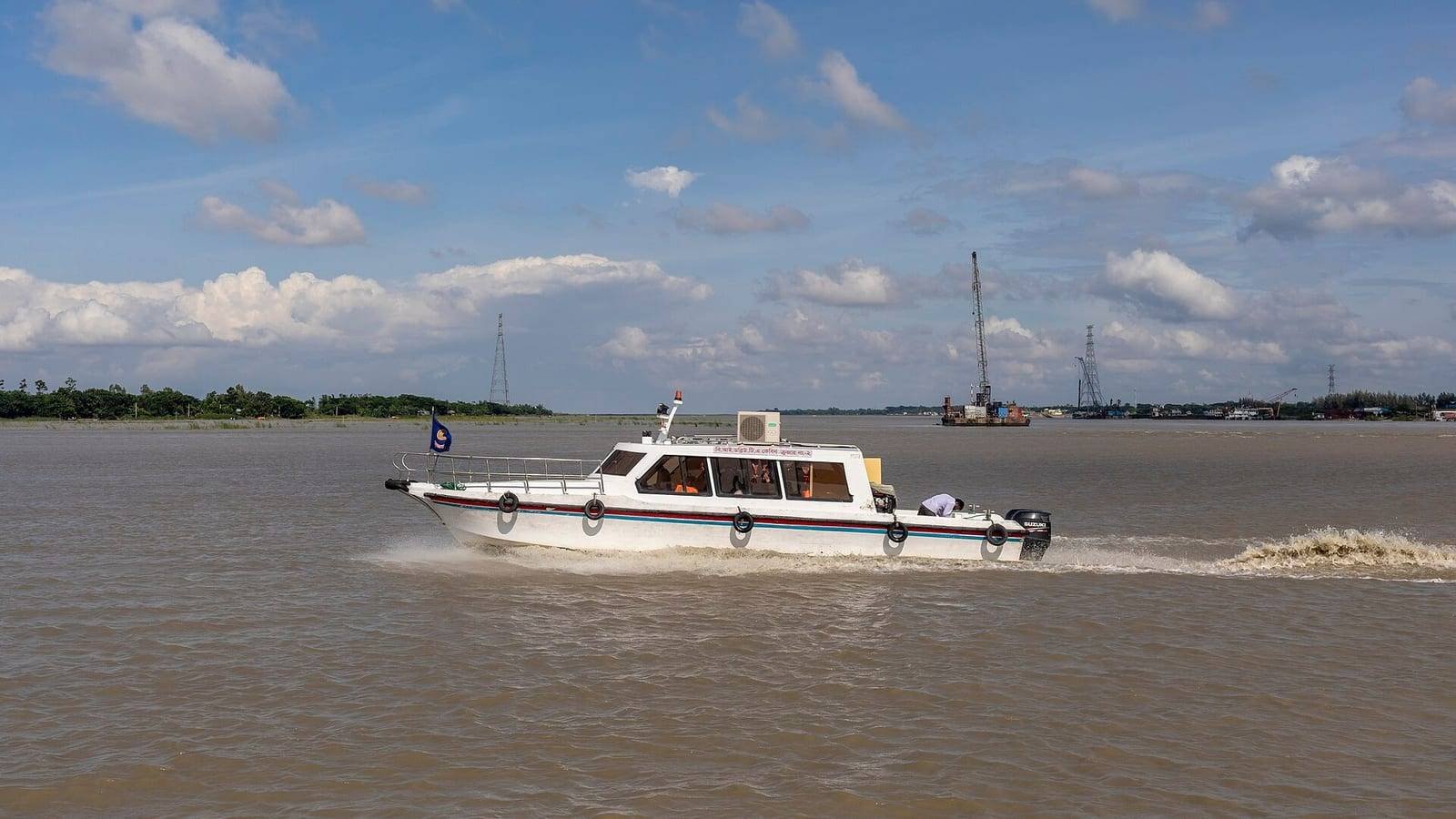 A white cabin speedboat running fast across a wide, silt-brown river under a blue sky, throwing spray astern, with low green banks and a crane on the far shore.