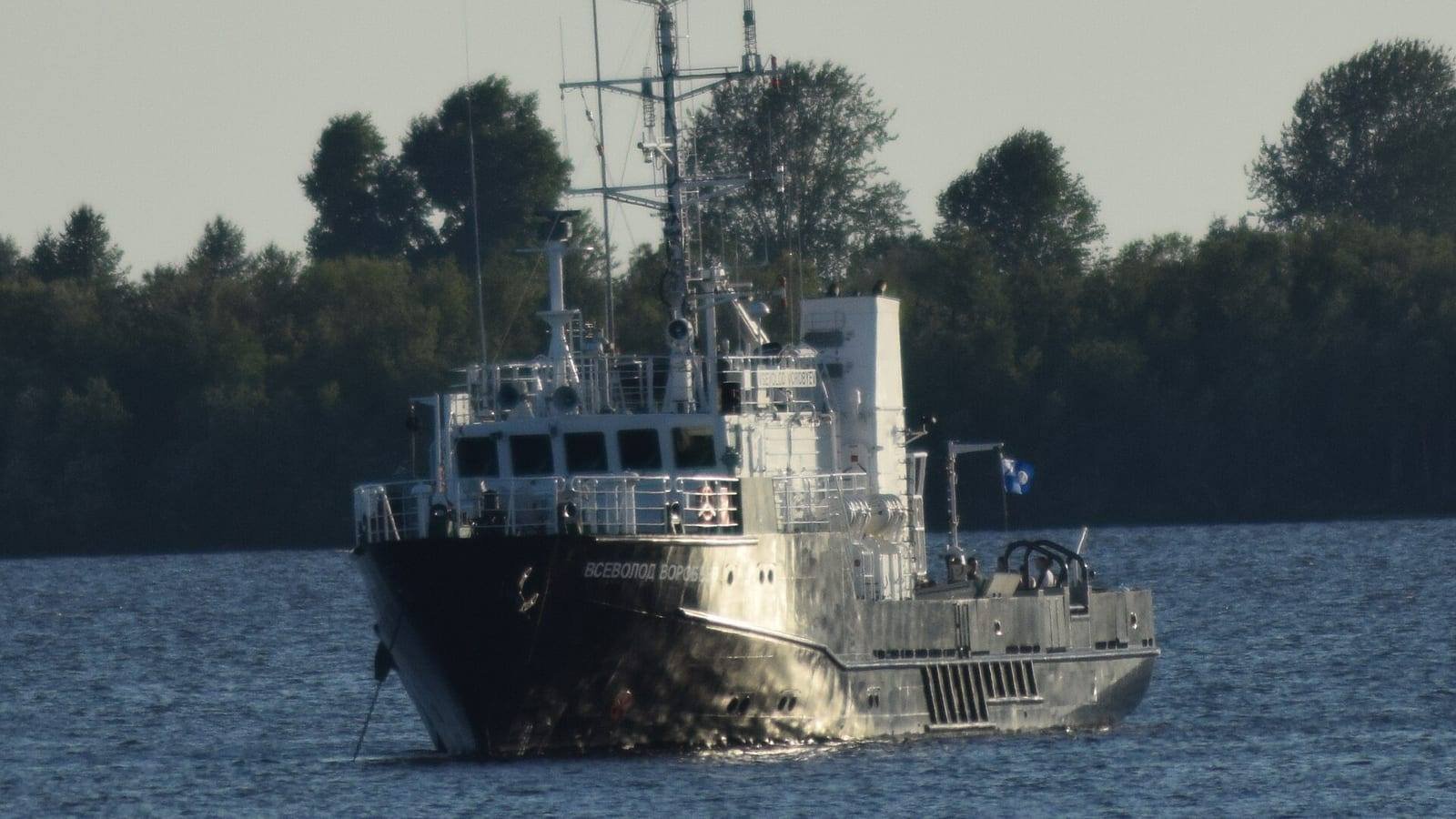 A grey steel survey vessel under way on a calm river, her mast carrying aerials and a radar above a low wheelhouse, with a wooded bank behind.